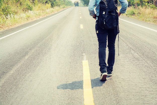 Rear view of a young woman hitchhiking carrying backpack walking on the road