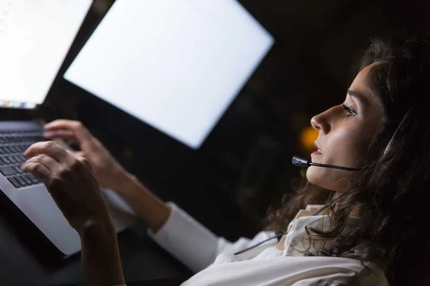 Businesswoman in headset using laptop. Side view of young serious call center operator working with laptop computer in dark office. Client service concept