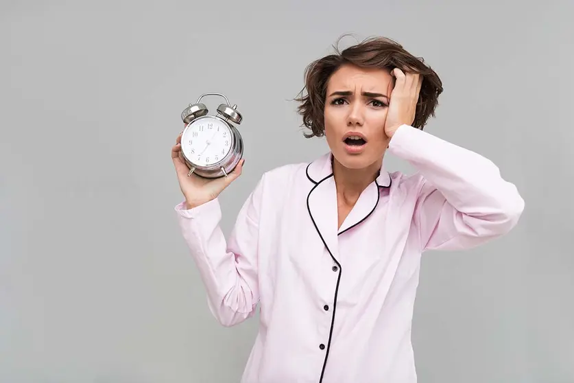 Portrait of a young confused girl in pajamas holding alarm clock and looking at camera isolated over gray background