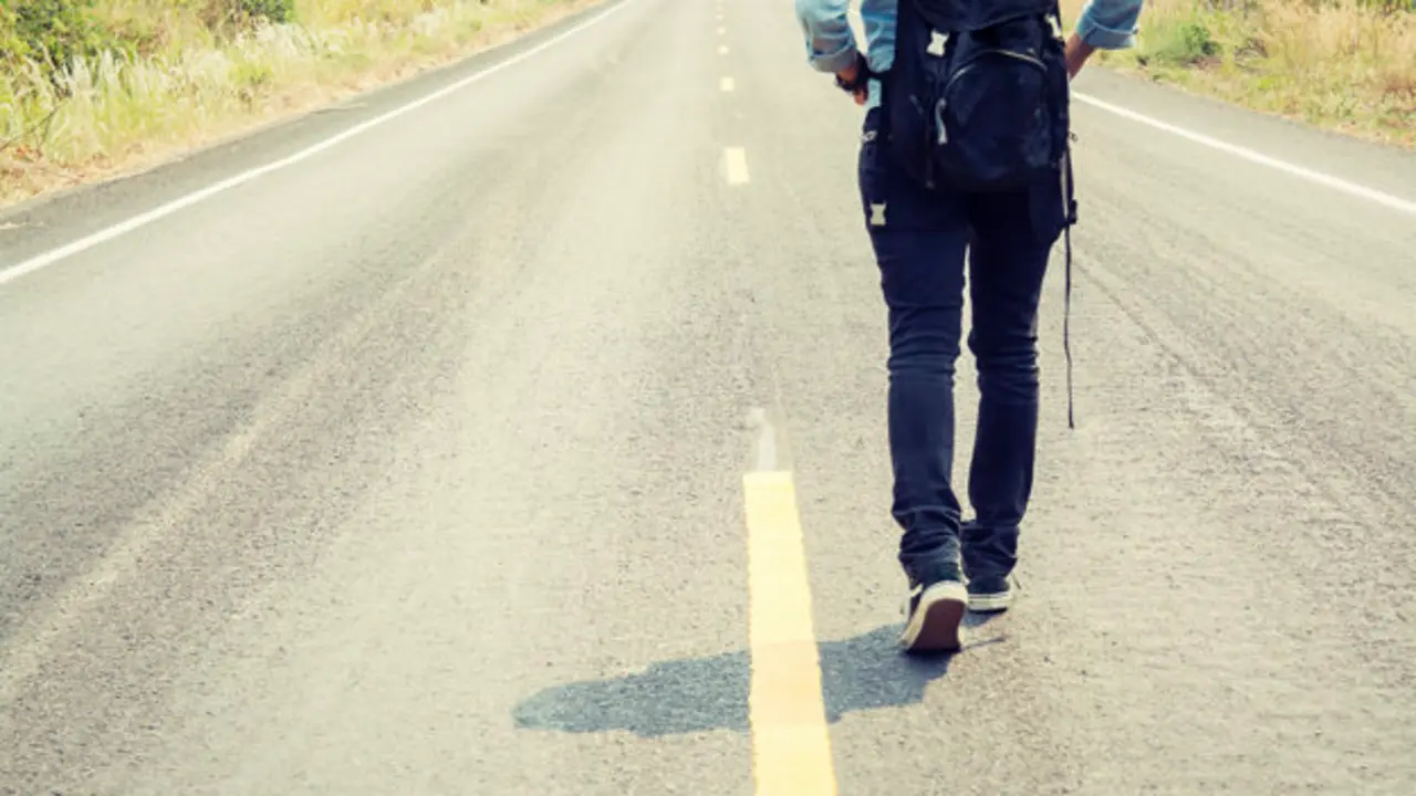 Rear view of a young woman hitchhiking carrying backpack walking on the road