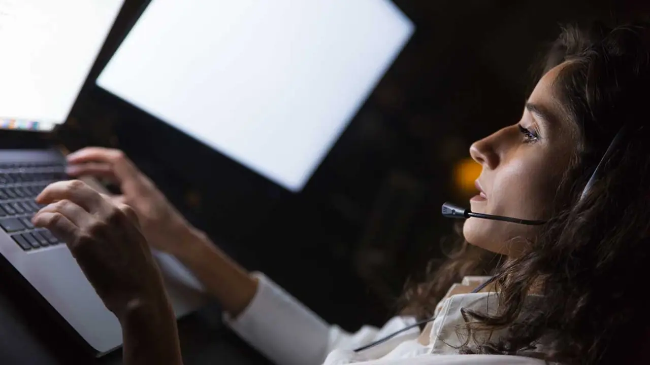 Businesswoman in headset using laptop. Side view of young serious call center operator working with laptop computer in dark office. Client service concept