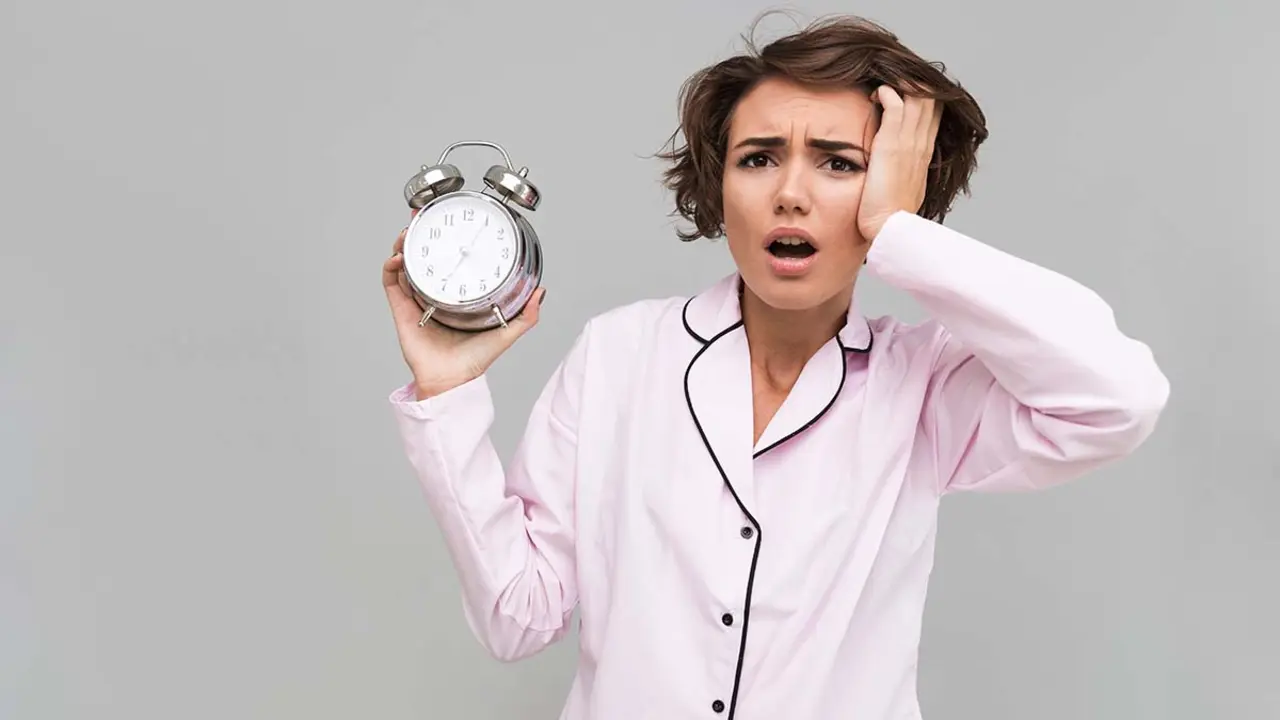 Portrait of a young confused girl in pajamas holding alarm clock and looking at camera isolated over gray background