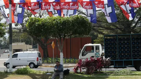 Un hombre almuerza debajo de la sombra de un árbol. Arriba de él banderas de las principales fuerzas políticas de El Salvador