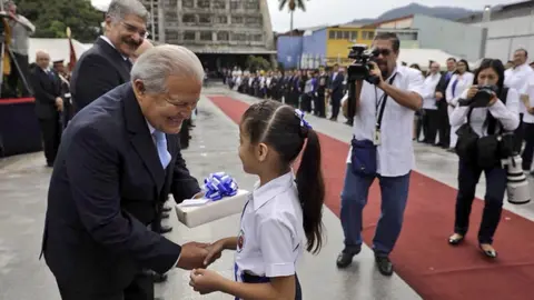 El presidente de la República, Salvador Sánchez Cerén, entrega un presente a la estudiante de primer grado que rezó la oración a la bandera en el acto cívico en la Plaza Libertad. Foto: Capres
Foto: Capres