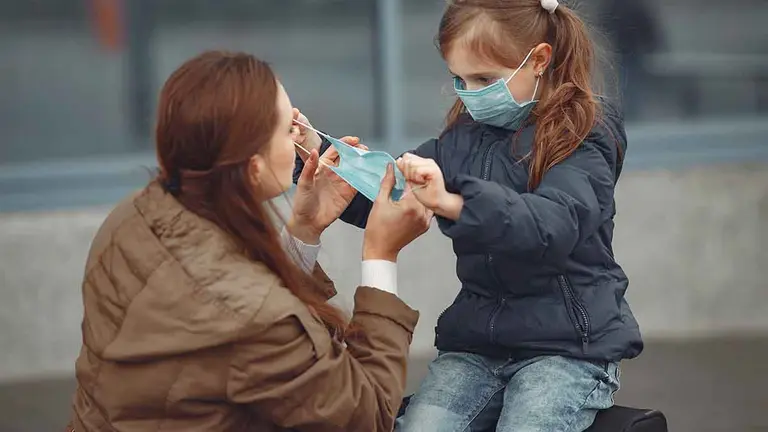 A European mother in a respirator with her daughter are standing near a building.The parent is teaching her child how to wear protective mask to save herself from virus.